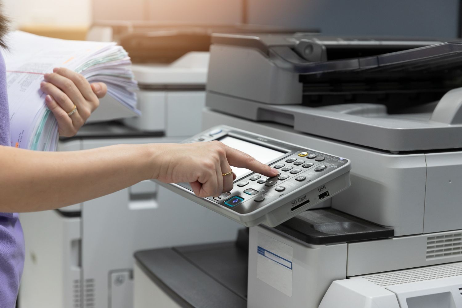 Person holding a stack of papers while pressing buttons on a photocopy machine.