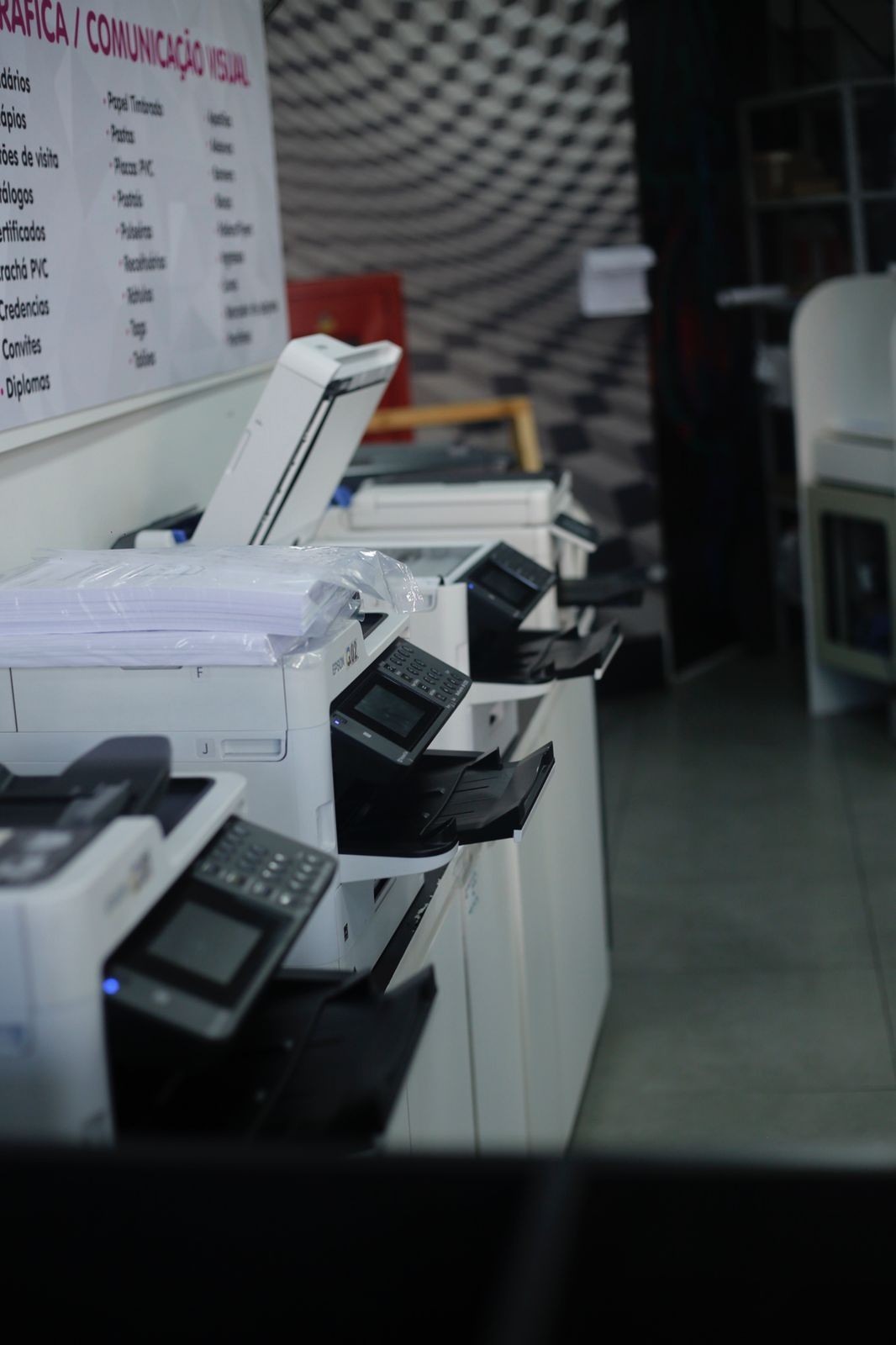 Row of modern printers in an office setting with a sign listing various printing services on the wall.